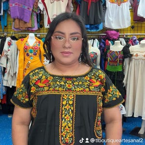 May include: A black embroidered top with short sleeves. The top features colorful floral and bird designs in yellow, red, and green. The background shows a clothing store with various embroidered garments.