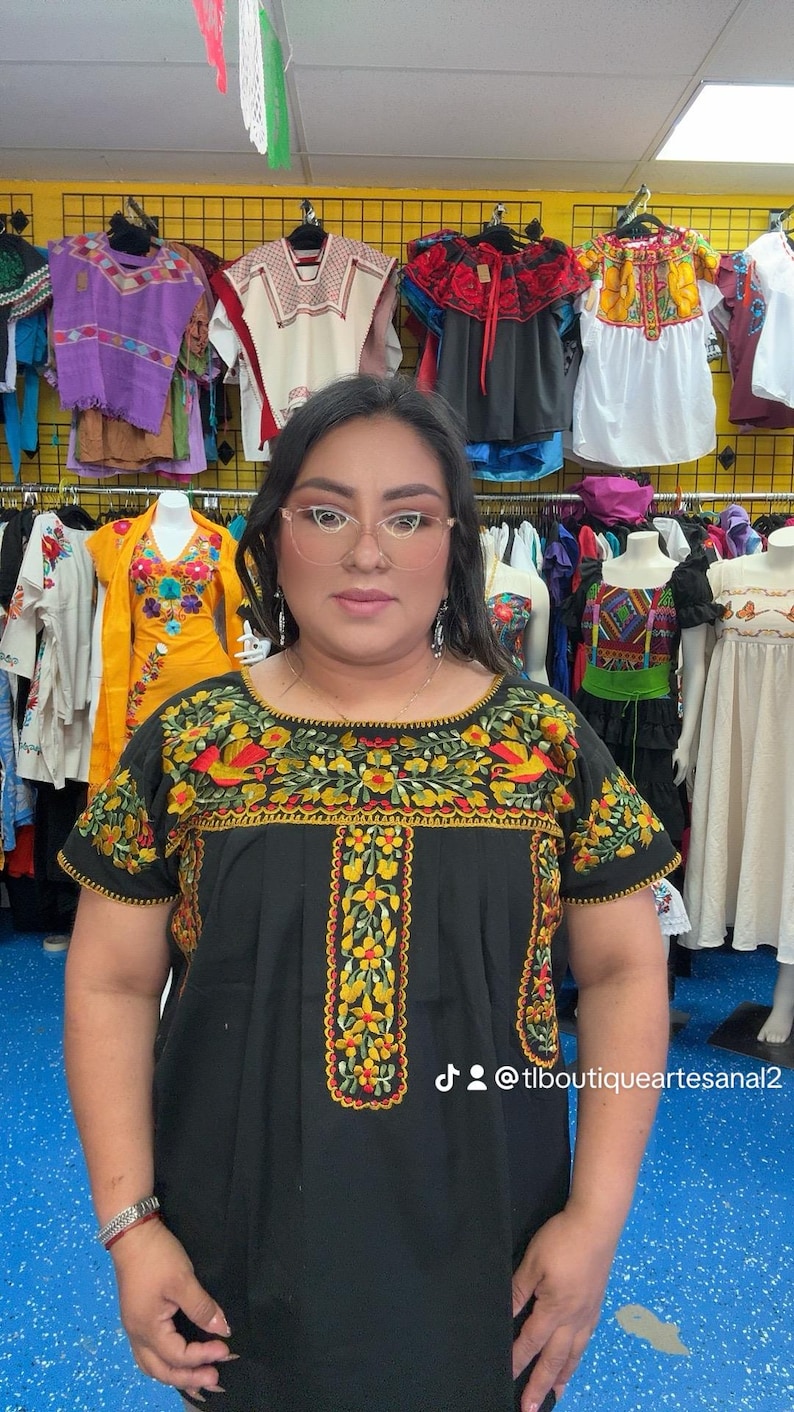 May include: A black embroidered top with short sleeves. The top features colorful floral and bird designs in yellow, red, and green. The background shows a clothing store with various embroidered garments.