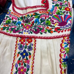 May include: White embroidered dress with vibrant floral patterns in red, pink, blue, green, and purple. The dress features a red trim and intricate stitching, showcasing a traditional design. The dress is made of a soft, flowing fabric.