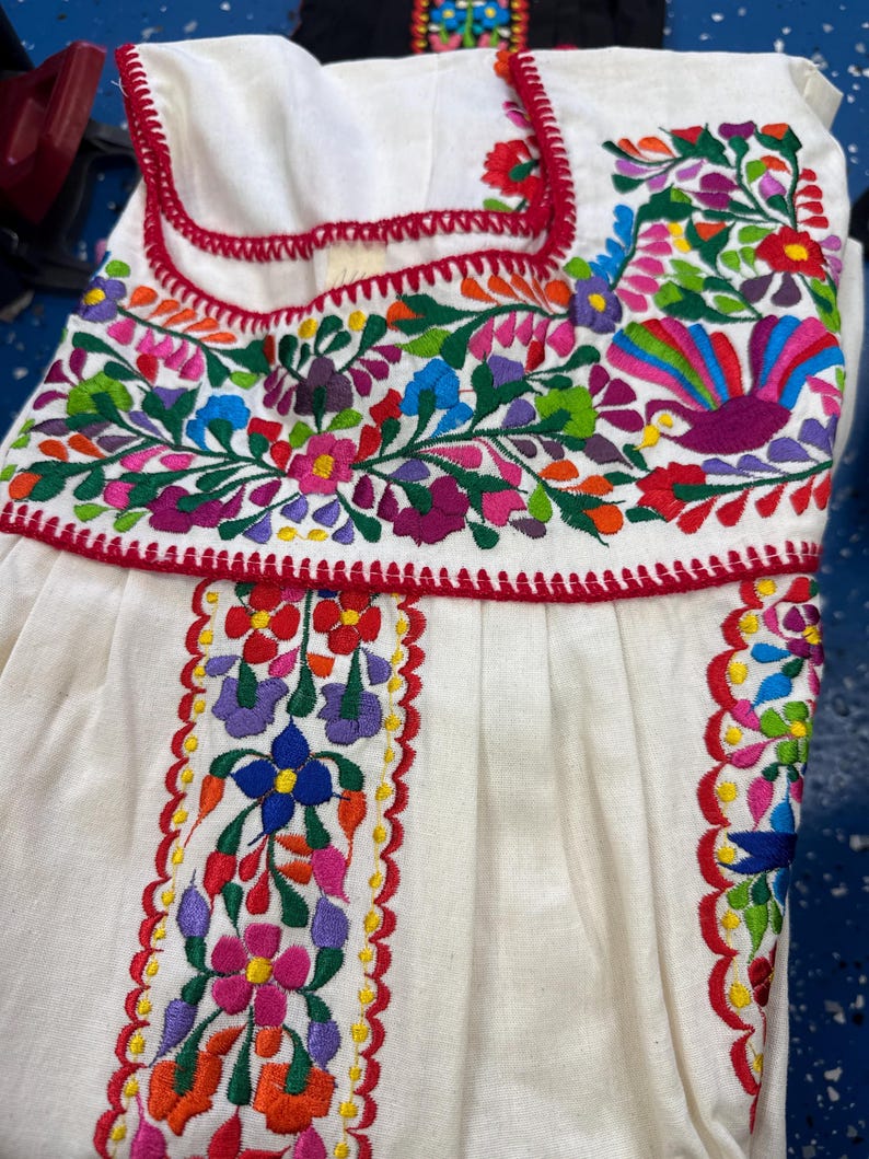 May include: White embroidered dress with vibrant floral patterns in red, pink, blue, green, and purple. The dress features a red trim and intricate stitching, showcasing a traditional design. The dress is made of a soft, flowing fabric.