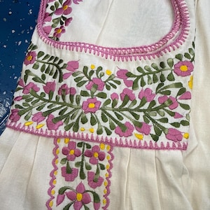 May include: White garment with intricate floral embroidery in pink, green, and yellow. The embroidery adorns the neckline and a vertical panel, creating a detailed and colorful design. The garment appears to be made of a soft, textured fabric.