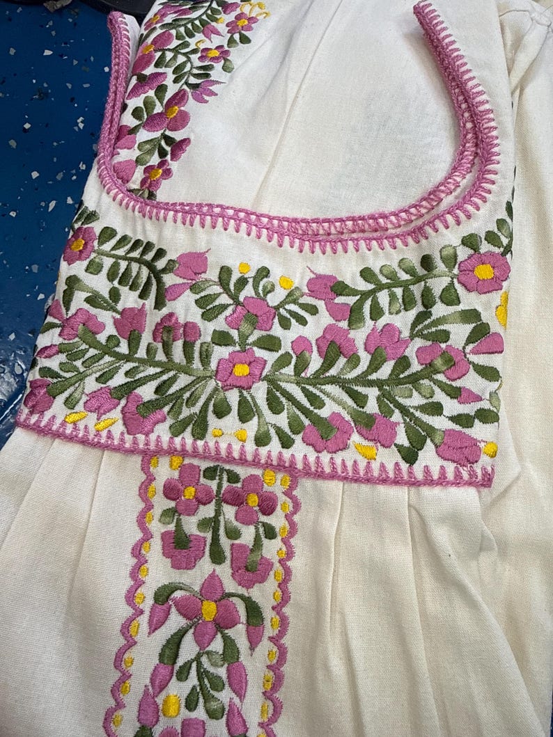 May include: White garment with intricate floral embroidery in pink, green, and yellow. The embroidery adorns the neckline and a vertical panel, creating a detailed and colorful design. The garment appears to be made of a soft, textured fabric.