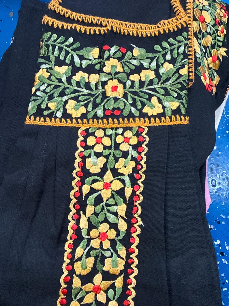 May include: Black embroidered top with floral designs. The embroidery features yellow flowers, green leaves, and red accents. The top has a square neckline and short sleeves, with a decorative trim.