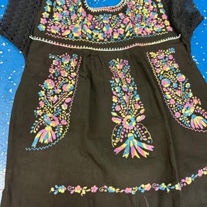 May include: Black embroidered tunic with colorful floral designs. The tunic features short black lace sleeves and intricate embroidery in shades of pink, yellow, blue, and purple. The neckline and hem are also adorned with floral embroidery.