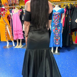 Pode incluir: Um vestido longo de cetim preto com decote ombro a ombro, mangas curtas e saia evas&ecirc; estilo sereia. A pe&ccedil;a possui um corpete justo que transita para uma bainha volumosa e fluida com um acabamento liso e reflexivo.