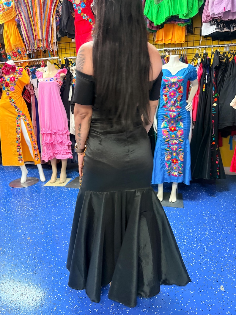 Pode incluir: Um vestido longo de cetim preto com decote ombro a ombro, mangas curtas e saia evas&ecirc; estilo sereia. A pe&ccedil;a possui um corpete justo que transita para uma bainha volumosa e fluida com um acabamento liso e reflexivo.