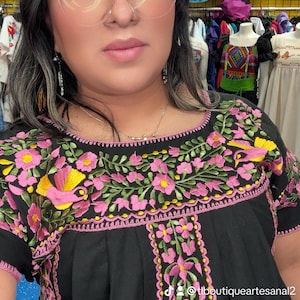 May include: Black embroidered top with pink and yellow floral design. The top features short sleeves and a round neckline with detailed embroidery. The design includes a central vertical floral pattern and a decorative border. The text "@tlboutiqueartezanal2" is visible.