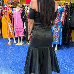 Pode incluir: Um vestido preto longo at&eacute; ao ch&atilde;o com decote ombro a ombro e mangas curtas. A pe&ccedil;a apresenta um corpete justo que transita para uma saia larga estilo sereia com uma bainha ampla e fluida feita de um tecido liso semelhante a cetim.