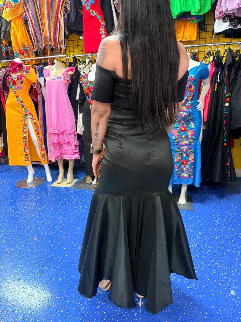 Pode incluir: Um vestido preto longo at&eacute; ao ch&atilde;o com decote ombro a ombro e mangas curtas. A pe&ccedil;a apresenta um corpete justo que transita para uma saia larga estilo sereia com uma bainha ampla e fluida feita de um tecido liso semelhante a cetim.