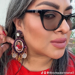 May include: A close-up of a purple and gold beaded earring featuring a portrait of Frida Kahlo. The earring has a large, oval-shaped design with a central image and dangling accents. The image is framed by a beaded border and a large crystal at the top.