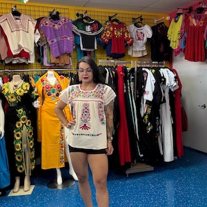 May include: A woman wearing a white embroidered top with colorful floral designs and black shorts. The image showcases a variety of traditional Mexican clothing, including blouses and dresses, displayed on racks and mannequins in a store setting.