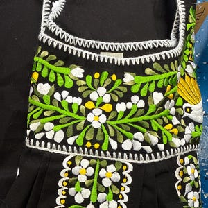 May include: Black garment with white, green, and yellow floral embroidery. The embroidery features detailed white flowers with yellow centers, green leaves, and a white scalloped trim. The garment has a square neckline and a pleated skirt.