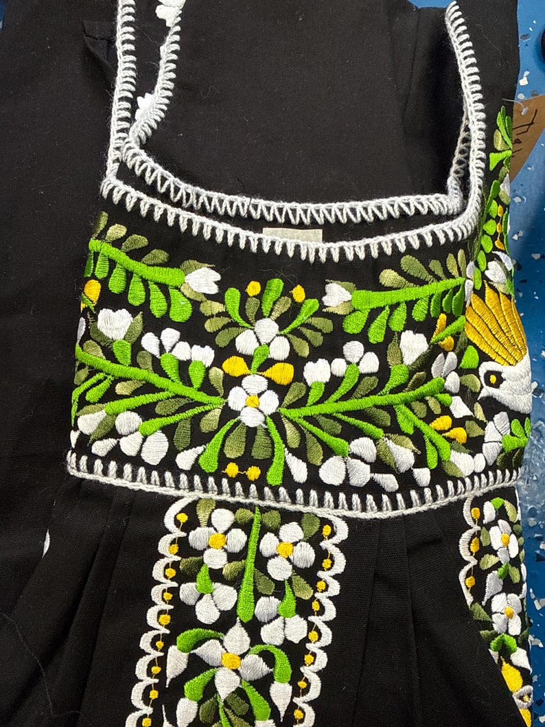 May include: Black garment with white, green, and yellow floral embroidery. The embroidery features detailed white flowers with yellow centers, green leaves, and a white scalloped trim. The garment has a square neckline and a pleated skirt.