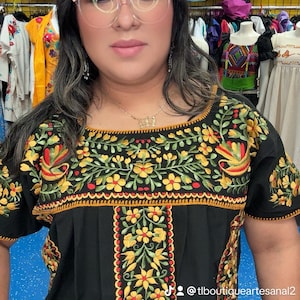 May include: Black embroidered top with yellow and red floral designs. The top features short sleeves and a square neckline. The embroidery is detailed with green leaves and a scalloped edge. The background includes other colorful clothing items.