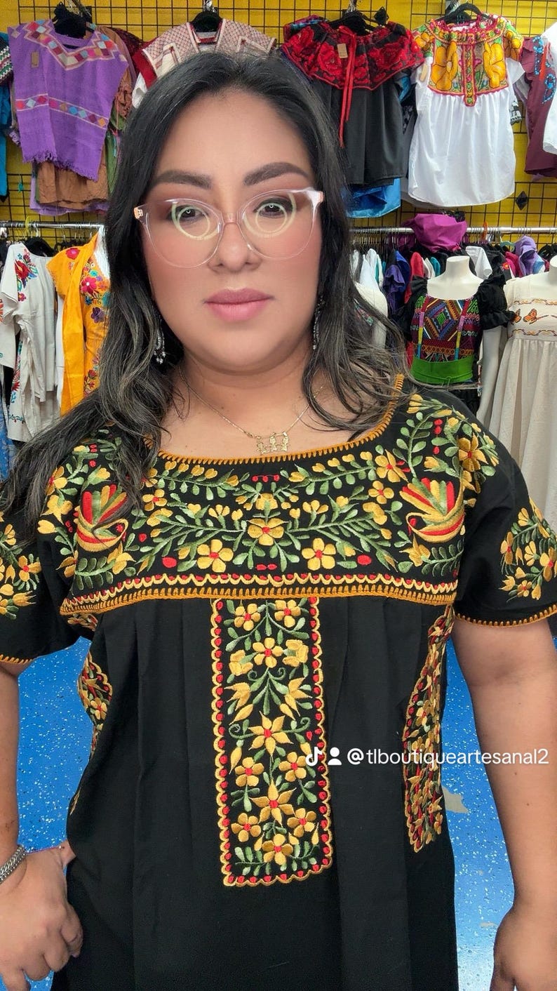 May include: Black embroidered top with yellow and red floral designs. The top features short sleeves and a square neckline. The embroidery is detailed with green leaves and a scalloped edge. The background includes other colorful clothing items.