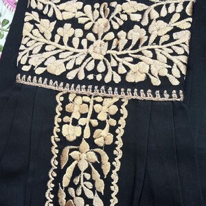 May include: Black garment with intricate beige floral embroidery. The embroidery is detailed with a scalloped edge along the neckline and vertical panel. The design features a central floral motif and symmetrical patterns, creating a classic and elegant look.