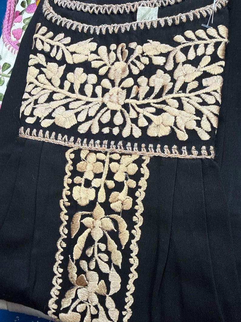 May include: Black garment with intricate beige floral embroidery. The embroidery is detailed with a scalloped edge along the neckline and vertical panel. The design features a central floral motif and symmetrical patterns, creating a classic and elegant look.
