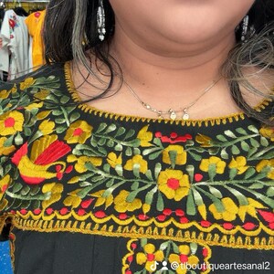 May include: Black embroidered top with vibrant floral patterns in yellow, red, and green. The design includes a square neckline and short sleeves. The text "@tlboutiqueartesanal2" is visible. The top is part of a clothing collection.