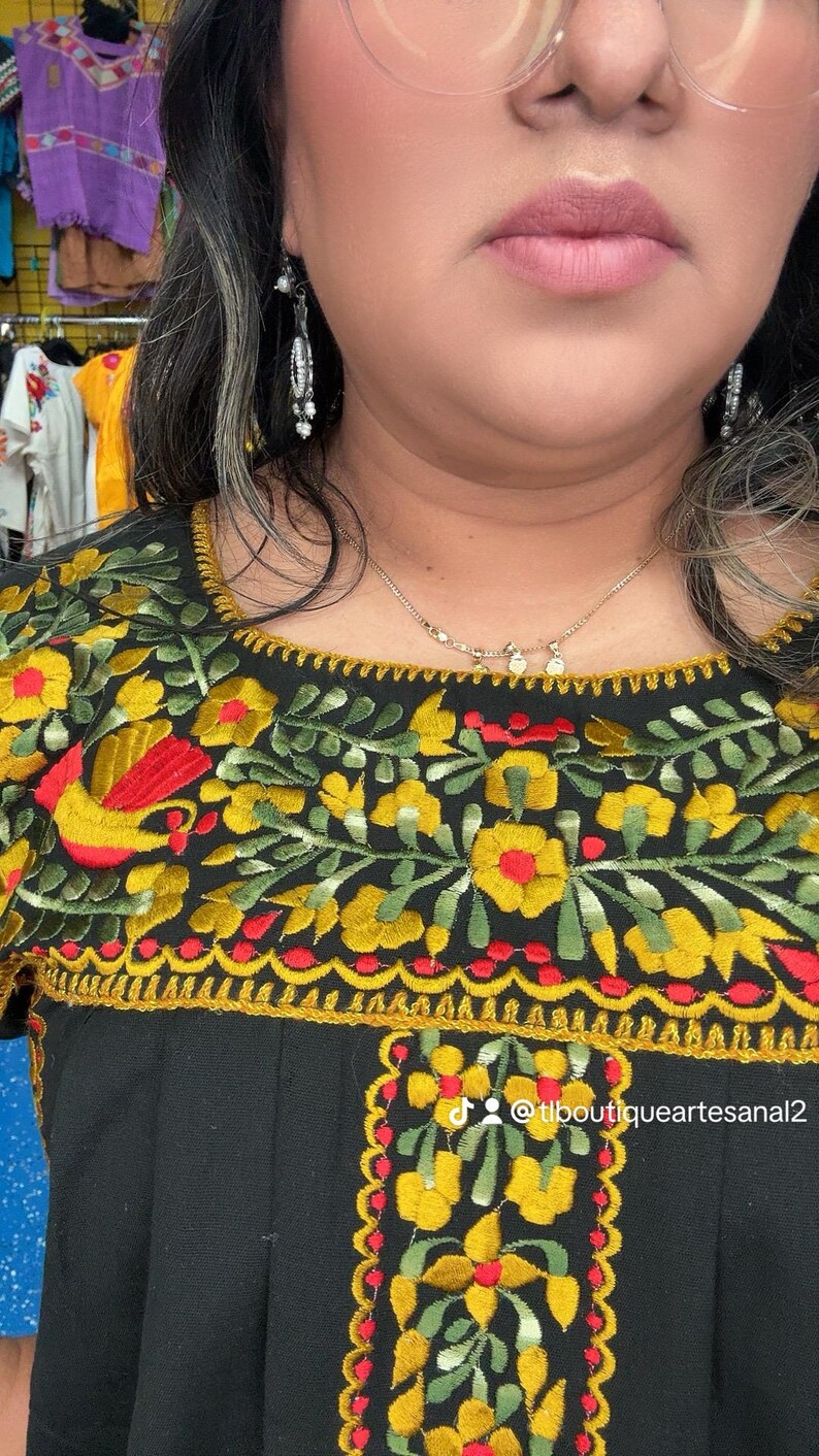 May include: Black embroidered top with vibrant floral patterns in yellow, red, and green. The design includes a square neckline and short sleeves. The text "@tlboutiqueartesanal2" is visible. The top is part of a clothing collection.