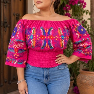 May include: A bright fuchsia off-the-shoulder blouse featuring intricate, multi-colored floral embroidery across the chest and sleeves. The garment has a smocked, elasticized waistline and three-quarter length sleeves, paired with blue denim jeans.