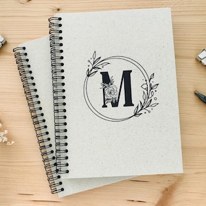 Könnte beinhalten: Zwei spiralgebundene Notizbücher mit weißem Einband und schwarzer Tinte. Das obere Notizbuch hat ein florales Design mit dem Buchstaben "M" in der Mitte.