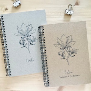 Könnte beinhalten: Zwei braune Spiralblock-Notizbücher mit schwarzer Wire-O-Bindung. Die Cover zeigen eine Schwarzweiß-Zeichnung einer Magnolienblüte. Das Notizbuch links trägt den Namen "Amelie". Das Notizbuch rechts trägt den Namen "Celina" und den Text "Notizen & Gedanken".