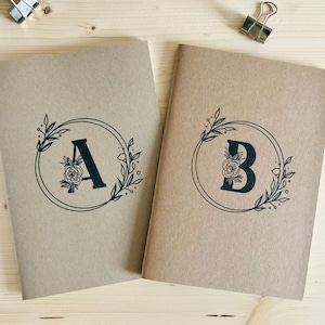 Könnte beinhalten: Zwei braune Kraftpapier-Notizbücher mit floralen Designs und den Buchstaben A und B in Schwarz.