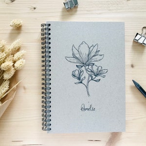 以下が含まれることがあります： 表紙に白黒のマグノリアの花の線画が描かれた茶色のスパイラルノート。ノートは木製の表面に置かれ、花の線画の下に「Amelie」という名前が筆記体で書かれている。