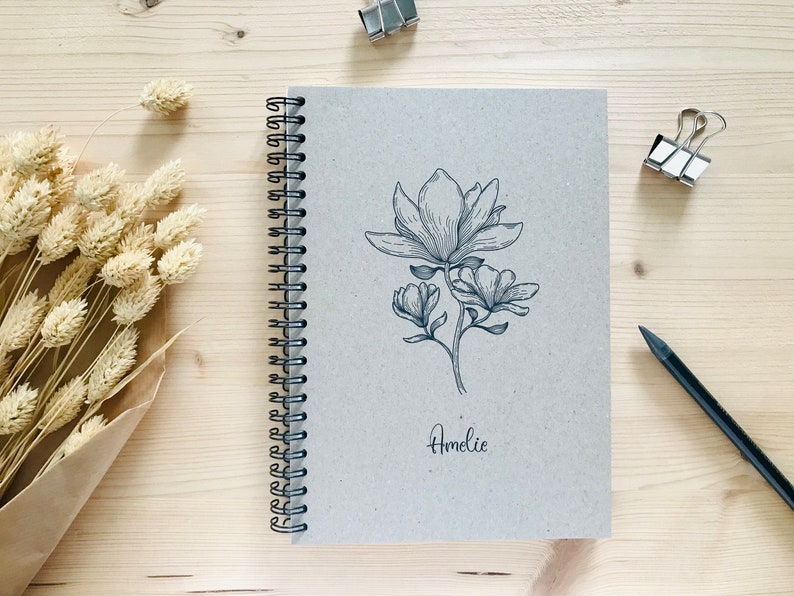 以下が含まれることがあります： 表紙に白黒のマグノリアの花の線画が描かれた茶色のスパイラルノート。ノートは木製の表面に置かれ、花の線画の下に「Amelie」という名前が筆記体で書かれている。