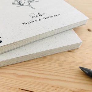 May include: Two spiral-bound notebooks with light-colored speckled covers are stacked on a wooden surface. The top notebook has a floral illustration and the text "Rebecca Notizen & Gedanken". A black pen is in the foreground.