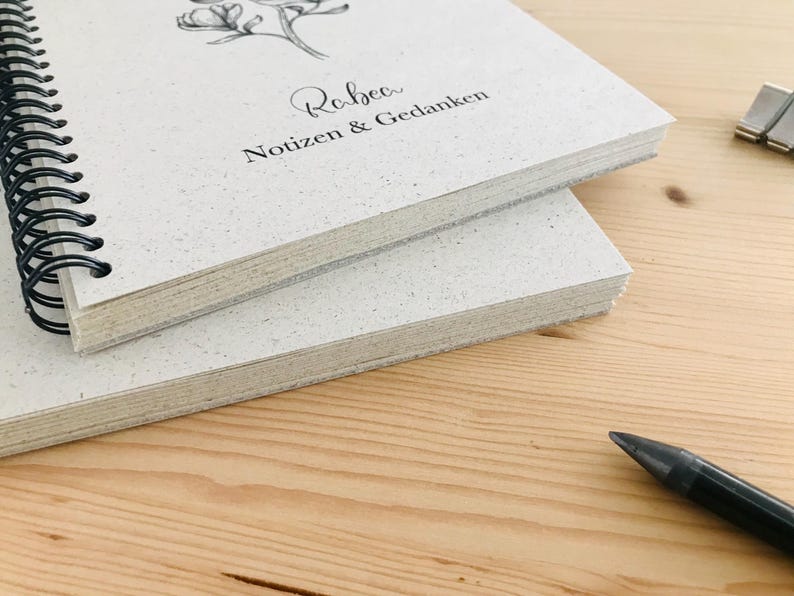 May include: Two spiral-bound notebooks with light-colored speckled covers are stacked on a wooden surface. The top notebook has a floral illustration and the text "Rebecca Notizen & Gedanken". A black pen is in the foreground.