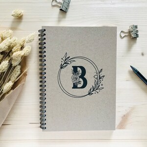 May include: A brown spiral bound notebook with a black floral design featuring the letter "B" in a circle. The notebook is on a light wood surface.