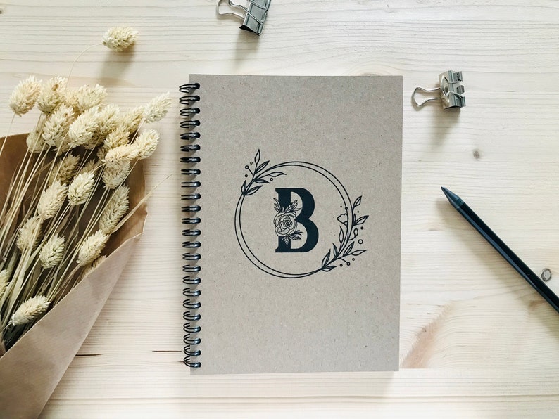 May include: A brown spiral bound notebook with a black floral design featuring the letter "B" in a circle. The notebook is on a light wood surface.