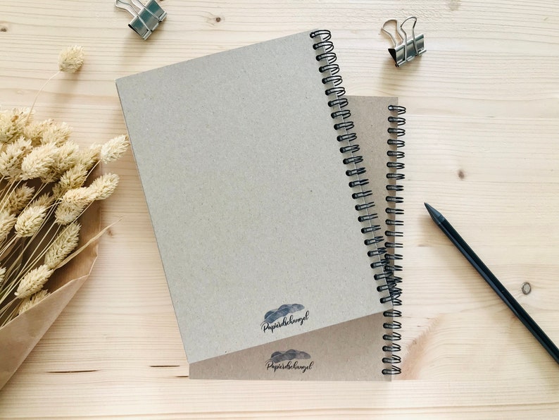 Puede incluir: Dos cuadernos de espiral marrones con el texto "papierdeengel" impreso en la portada. Los cuadernos est&aacute;n sobre una superficie de madera clara con un l&aacute;piz y clips.