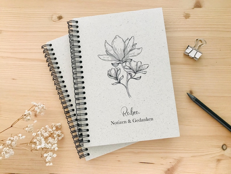 May include: Two spiral-bound notebooks with a floral illustration and the name "Rabea" and the text "Notizen & Gedanken". The notebooks are on a light wood surface, with a pencil and a binder clip.