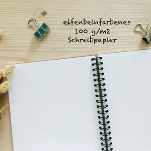 Puede incluir: Un cuaderno de espiral abierto con p&aacute;ginas blancas en blanco. El cuaderno est&aacute; sobre una superficie de madera con dos clips de carpeta plateados y tallos de flores secas. El texto "elfenbeinfarbenes 100 g/m2 Schreibpapier" est&aacute; escrito en la superficie.