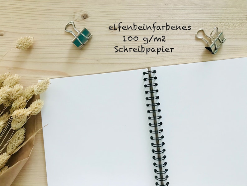 Puede incluir: Un cuaderno de espiral abierto con p&aacute;ginas blancas en blanco. El cuaderno est&aacute; sobre una superficie de madera con dos clips de carpeta plateados y tallos de flores secas. El texto "elfenbeinfarbenes 100 g/m2 Schreibpapier" est&aacute; escrito en la superficie.