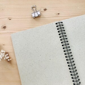 May include: An open, spiral-bound notebook with light gray speckled pages. A silver binder clip rests near the top, and a sprig of white flowers is in the lower left corner. The notebook is on a light wood surface.
