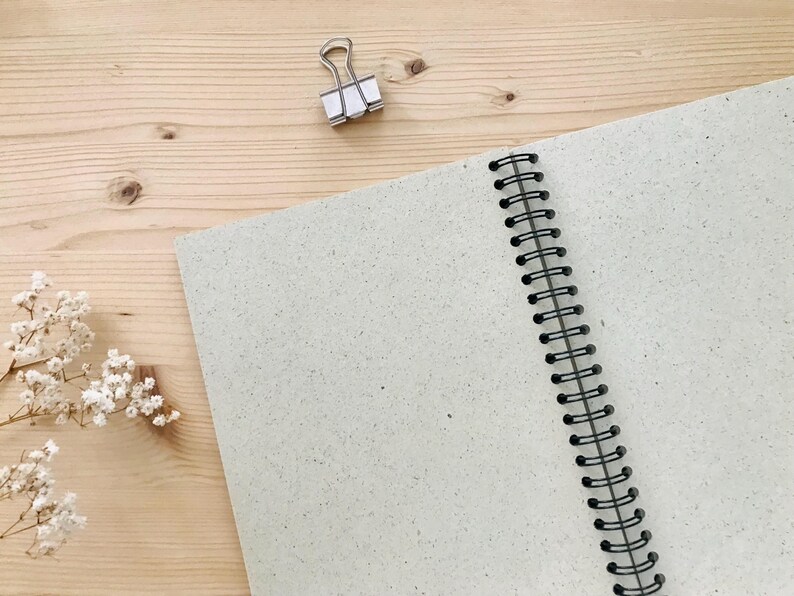 May include: An open, spiral-bound notebook with light gray speckled pages. A silver binder clip rests near the top, and a sprig of white flowers is in the lower left corner. The notebook is on a light wood surface.