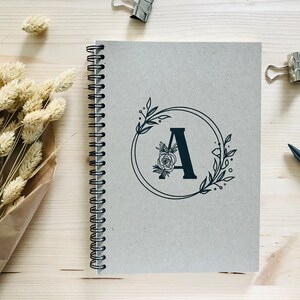 May include: A gray spiral bound notebook with a black floral design and the letter "A" in the center.