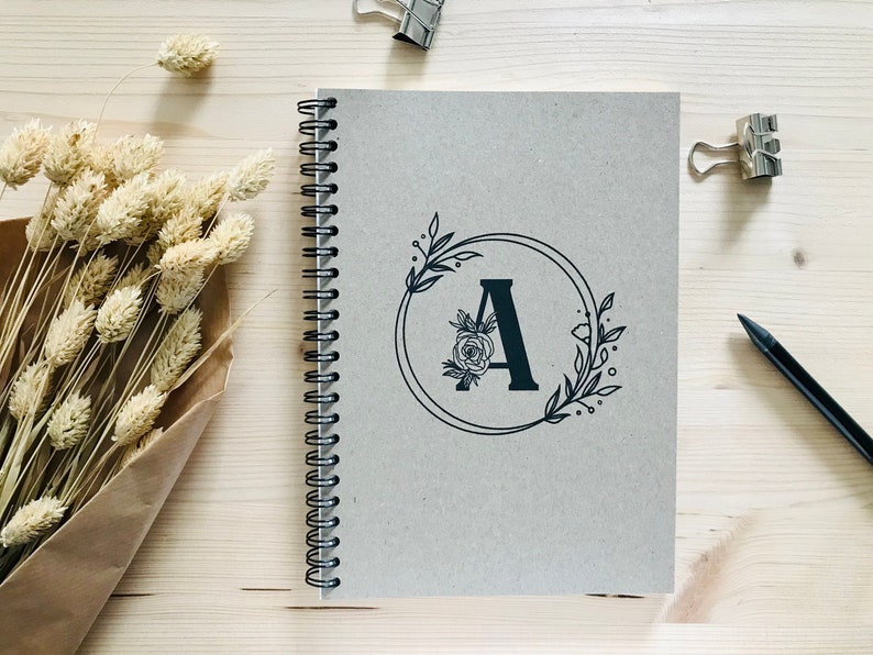 May include: A gray spiral bound notebook with a black floral design and the letter "A" in the center.