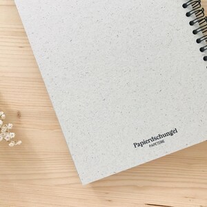 May include: A spiral-bound notebook with a textured, off-white cover, resting on a light wood surface. The notebook's black spiral binding is visible along the edge. The text "Papierschungel PAPETERIE" is printed on the cover.