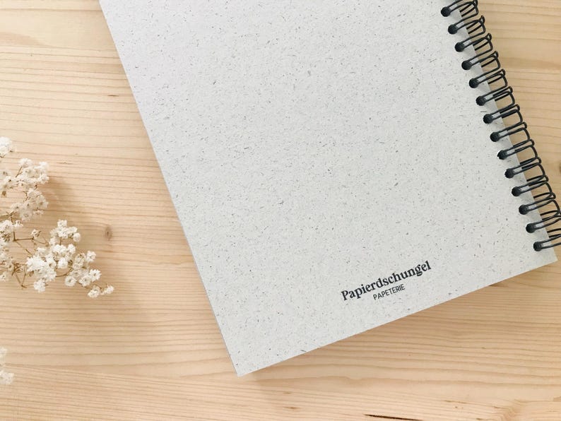 May include: A spiral-bound notebook with a textured, off-white cover, resting on a light wood surface. The notebook's black spiral binding is visible along the edge. The text "Papierschungel PAPETERIE" is printed on the cover.