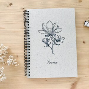 May include: A spiral-bound notebook with a floral illustration of a magnolia flower. The notebook has a light gray cover with the name "Bianca" written in cursive. A pen and binder clip are on the wooden surface.