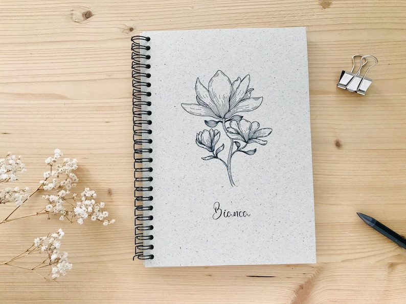 May include: A spiral-bound notebook with a floral illustration of a magnolia flower. The notebook has a light gray cover with the name "Bianca" written in cursive. A pen and binder clip are on the wooden surface.