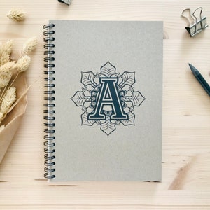 Puede incluir: Un cuaderno gris de espiral con un dise&ntilde;o de mandala en blanco y negro con la letra "A" en el centro.