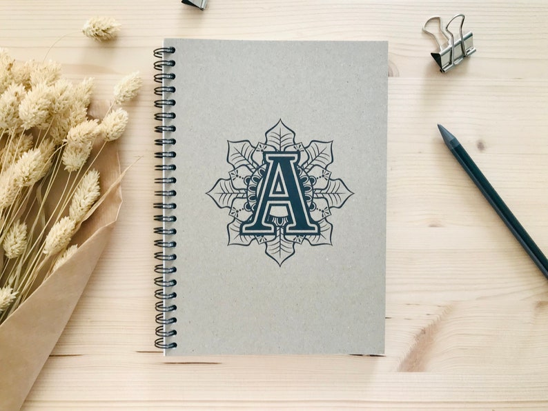 Puede incluir: Un cuaderno gris de espiral con un dise&ntilde;o de mandala en blanco y negro con la letra "A" en el centro.