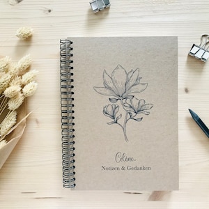 以下が含まれることがあります： マグノリアの花の白黒のイラストが描かれた茶色のスパイラルバインダーノート。ノートの表紙には「Celina Notizen & Gedanken」という文字が印刷されています。