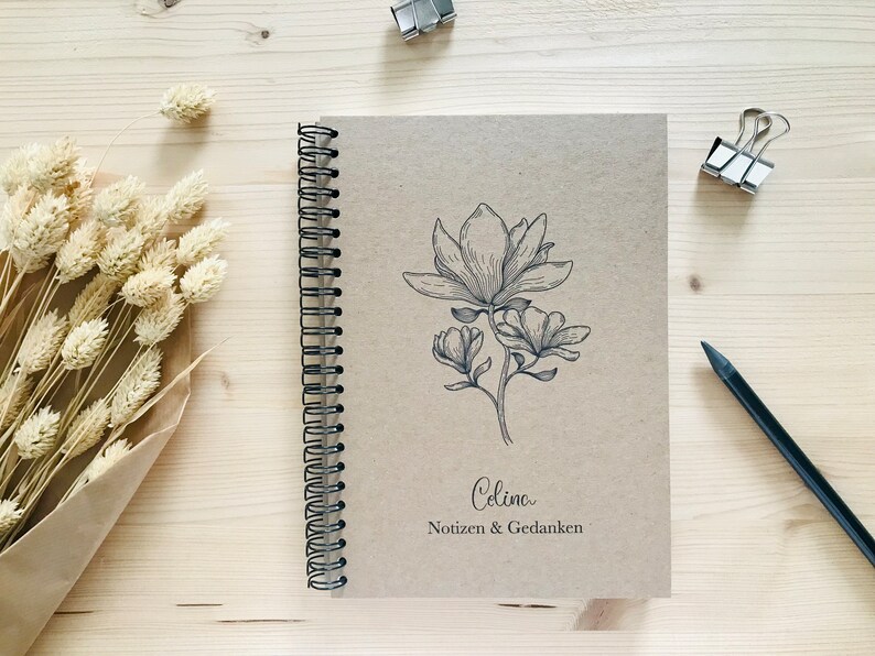 以下が含まれることがあります： マグノリアの花の白黒のイラストが描かれた茶色のスパイラルバインダーノート。ノートの表紙には「Celina Notizen & Gedanken」という文字が印刷されています。