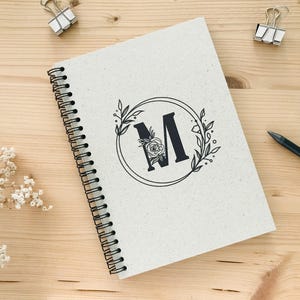 K&ouml;nnte beinhalten: Ein wei&szlig;es Spiralnotizbuch mit einem schwarzen floralen Design und dem Buchstaben "M" in der Mitte. Das Notizbuch hat eine schwarze Spiralbindung.